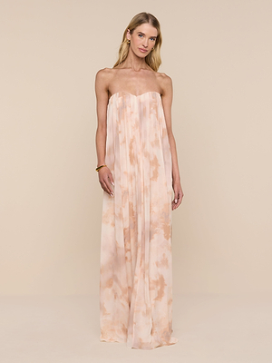 A woman with blonde hair and minimal jewelry stands against a beige backdrop wearing the Raine Strapless Gown, a floor-length dress featuring a built-in corset and loose, flowing fit with a light peach tie-dye pattern.