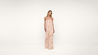 A woman stands in a strapless, floor-length, light pink and white patterned dress against a plain white background.