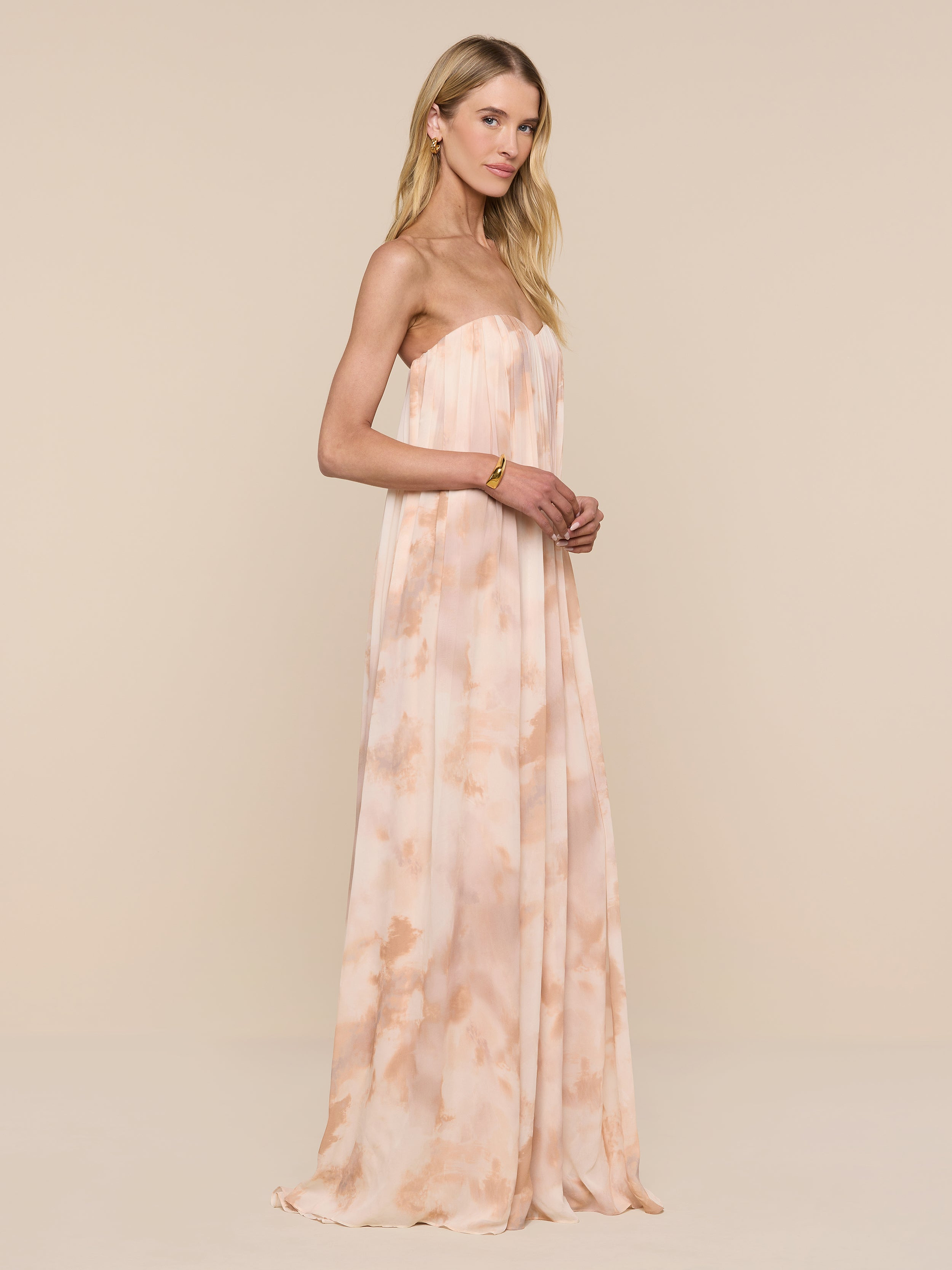 A woman poses against a plain white background in the Raine Strapless Gown, a floor-length dress with a peach and cream pattern. The gown features a built-in corset, and she wears her long blonde hair down with minimal accessories.