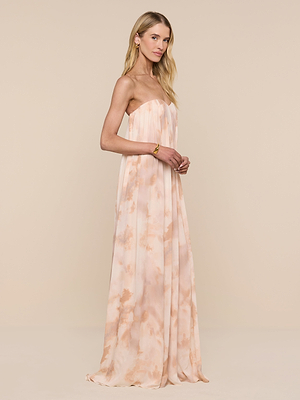A woman with long blonde hair wears the Raine Strapless Gown in peach and cream tie-dye, featuring a built-in corset. She stands sideways, smiling softly against a plain beige background.