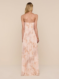 A woman with long blonde hair wears the Raine Strapless Gown, a floor-length dress in light peach featuring a subtle pattern and a built-in corset. She stands facing away against a plain white background.