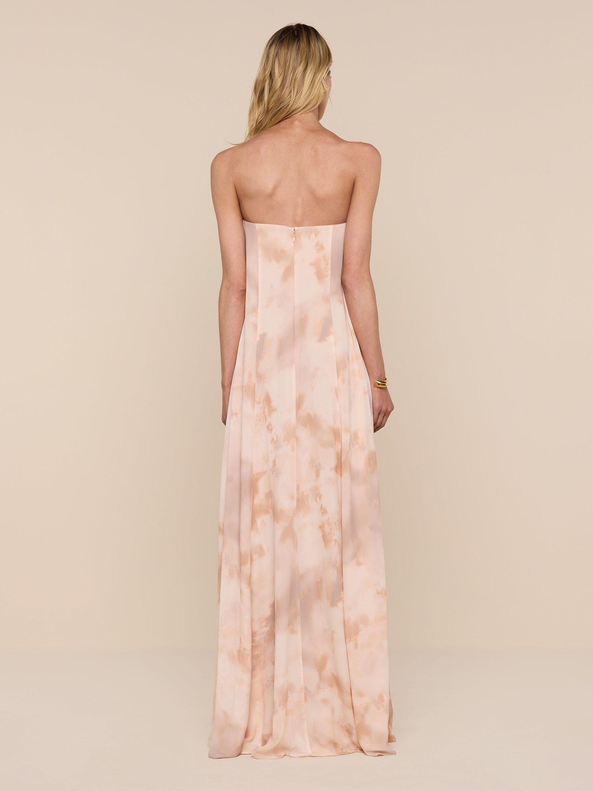 A woman with long blonde hair wears the Raine Strapless Gown, a floor-length dress in light peach featuring a subtle pattern and a built-in corset. She stands facing away against a plain white background.