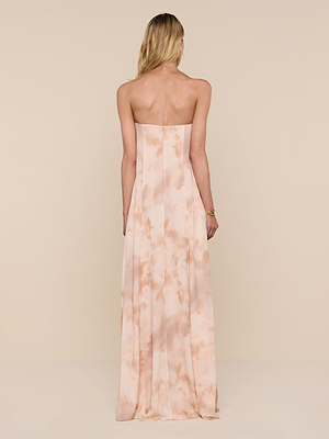 A woman with long blonde hair wears the Raine Strapless Gown—a floor-length peach and cream tie-dye dress with a built-in corset—as she stands facing away against a plain beige background, arms relaxed at her sides.