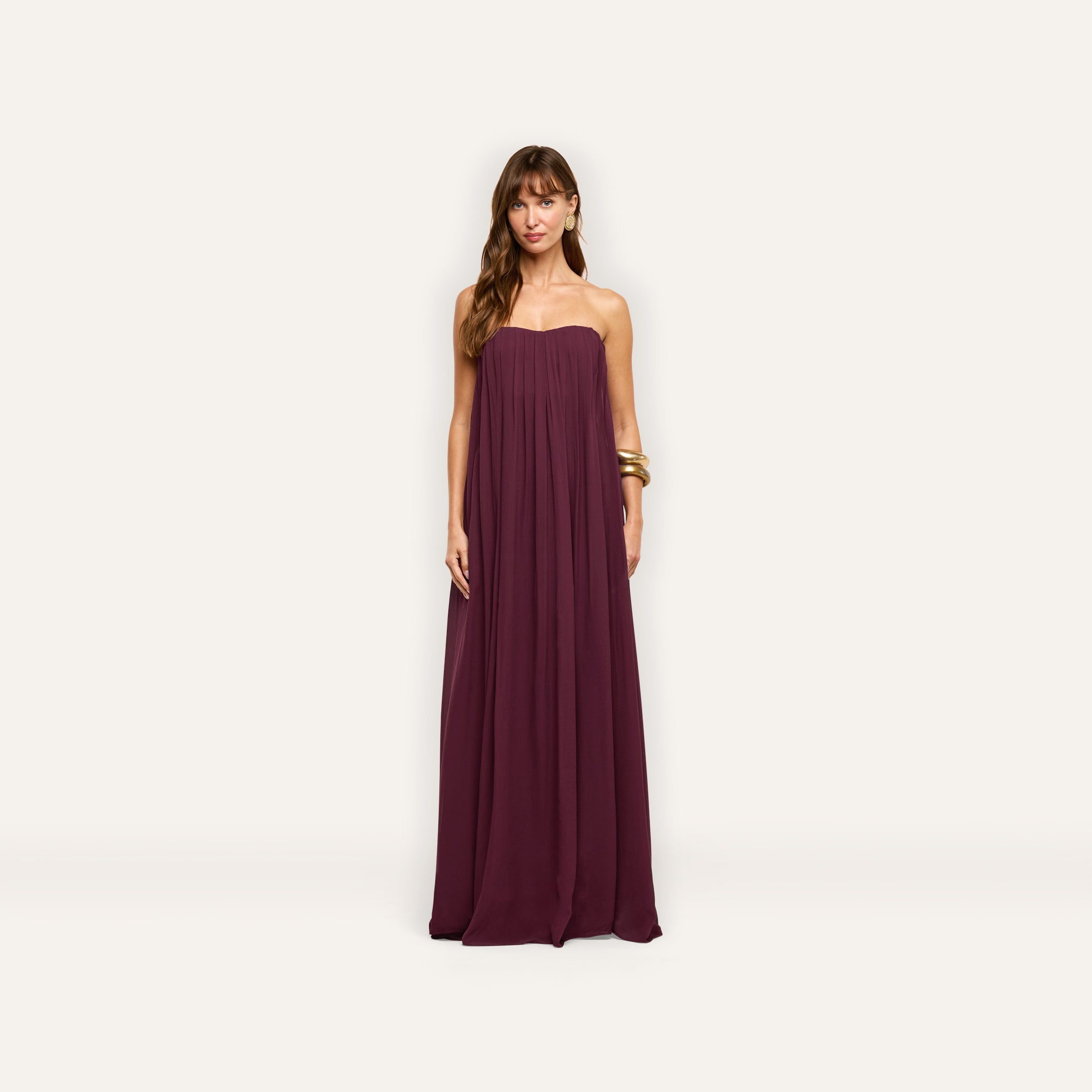 A woman models the floor-length, plum-colored Raine Strapless Gown, which features a strapless, pleated bodice made of viscose chiffon.