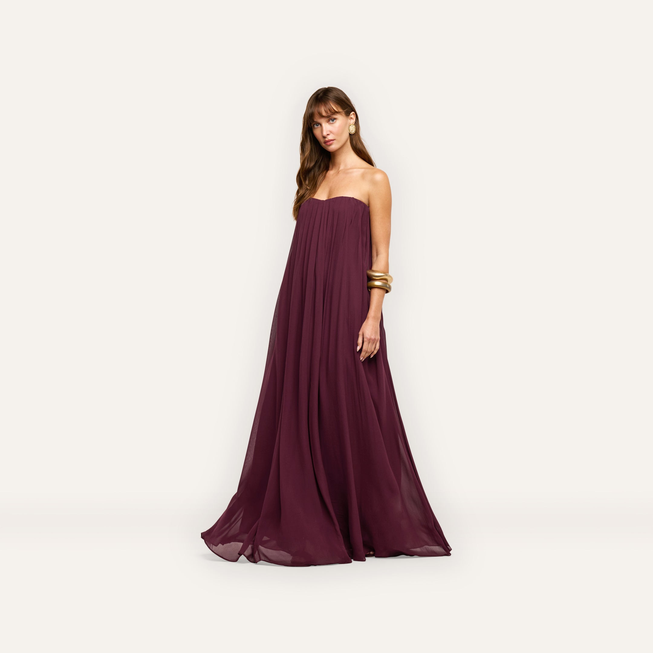 A person wearing a floor-length, strapless, pleated plum Raine Strapless Gown in viscose chiffon, accessorized with gold arm bangles.