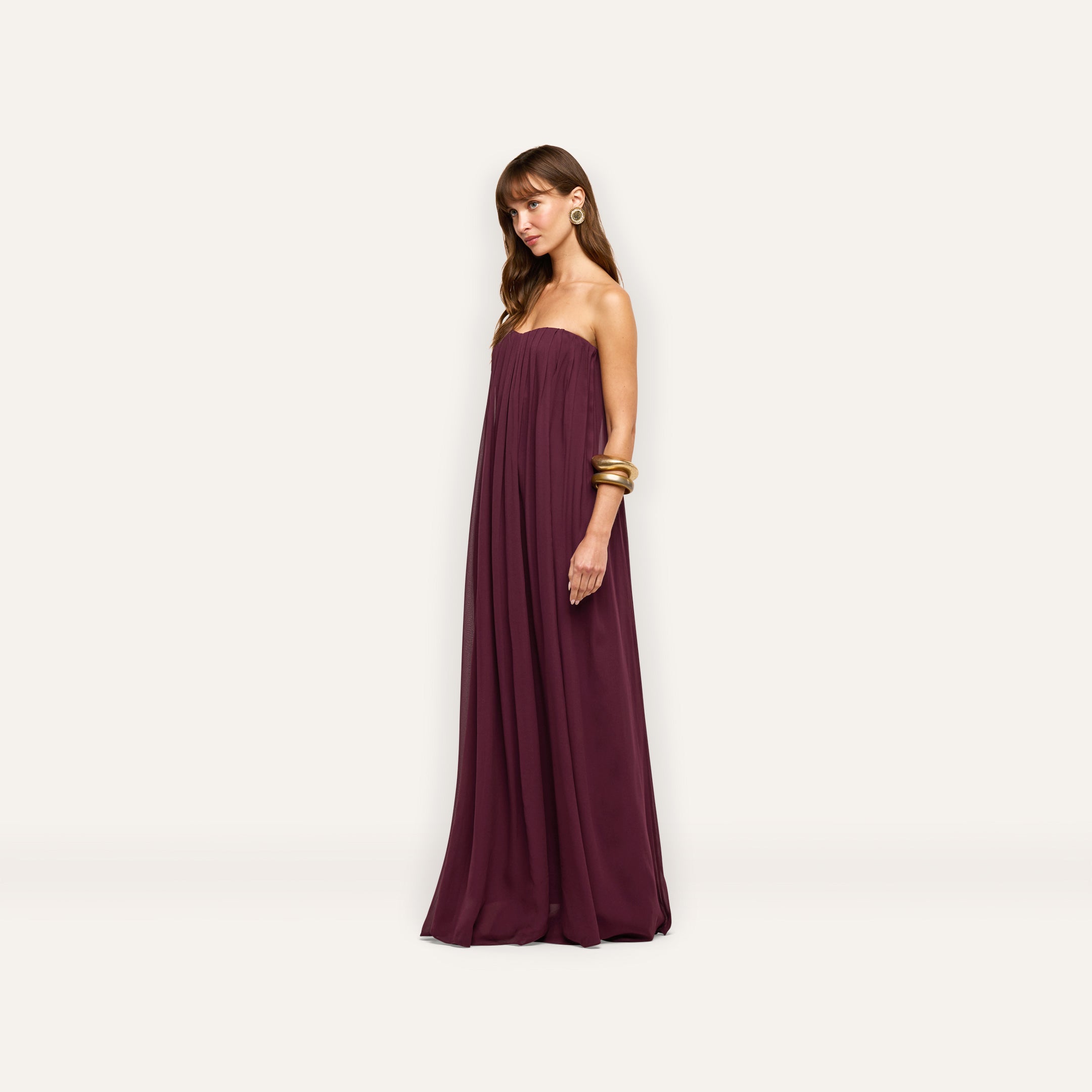 A woman wears a plum-colored Raine Strapless Gown in viscose chiffon, featuring a floor-length silhouette with a strapless bodice and elegant gathers.
