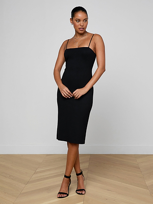 A woman poses with hands clasped, looking to the side, wearing the Eira Midi Dress—a fitted black slip dress with adjustable straps—paired with black heeled sandals. She stands on light wood flooring against a plain white wall.