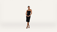Woman stands against a plain white background, wearing a black, sleeveless, knee-length dress and black high-heeled sandals. Her hands are clasped in front of her and she looks confidently at the camera.