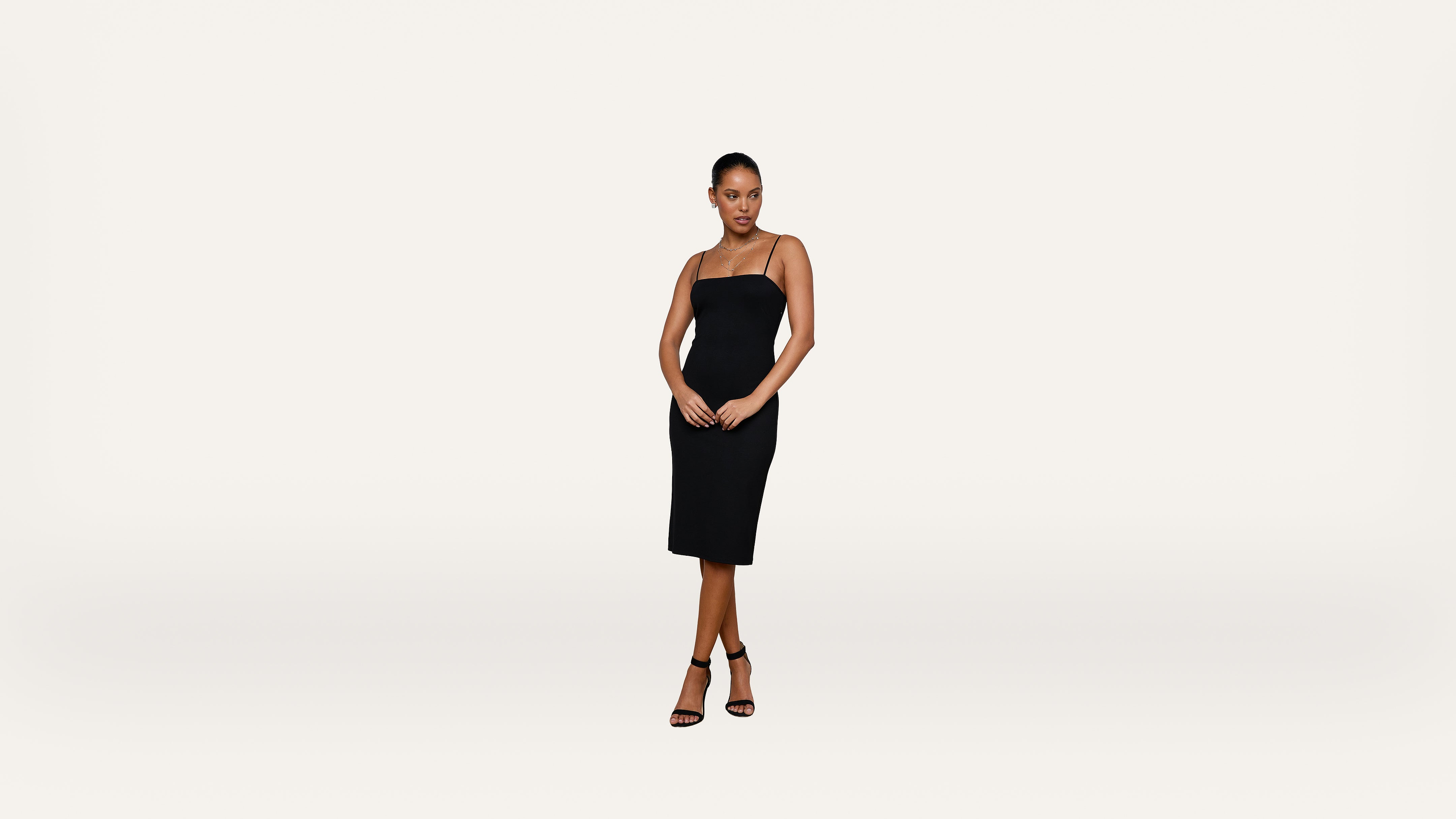 Woman stands against a plain white background, wearing a black, sleeveless, knee-length dress and black high-heeled sandals. Her hands are clasped in front of her and she looks confidently at the camera.