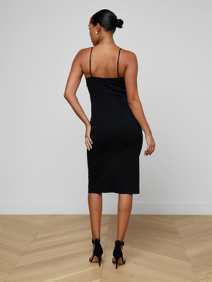 A woman with her hair in a bun stands with her back to the camera, wearing the fitted, sleeveless black Eira Midi Dress and black high-heeled shoes on a light wooden floor against a plain white wall.
