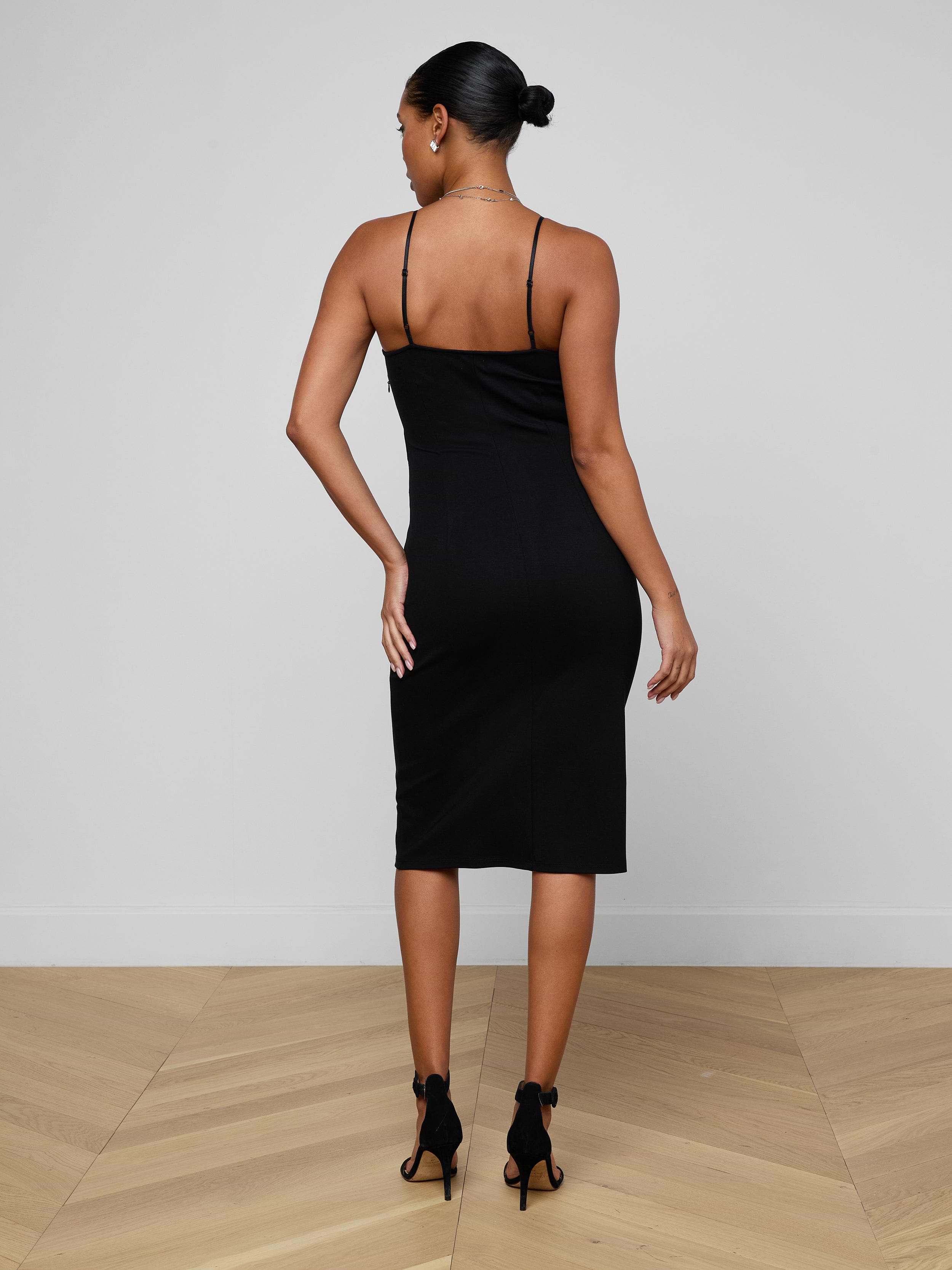 A woman with her hair in a bun wears the Eira Midi Dress—a fitted, sleeveless slip dress crafted from super stretch ponte—in black, paired with black heels. She stands with her back to the camera against a plain white background.