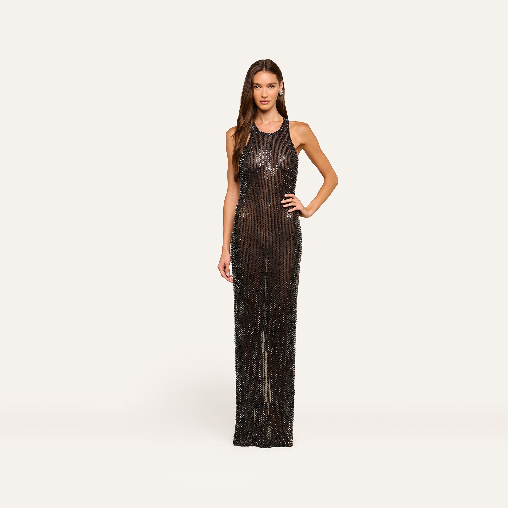 A woman stands confidently wearing the Paula Gown—a sleeveless, floor-length sheer mesh dress with a subtle shimmer. Her long hair is down, and she poses with one hand on her hip against a plain, light background.