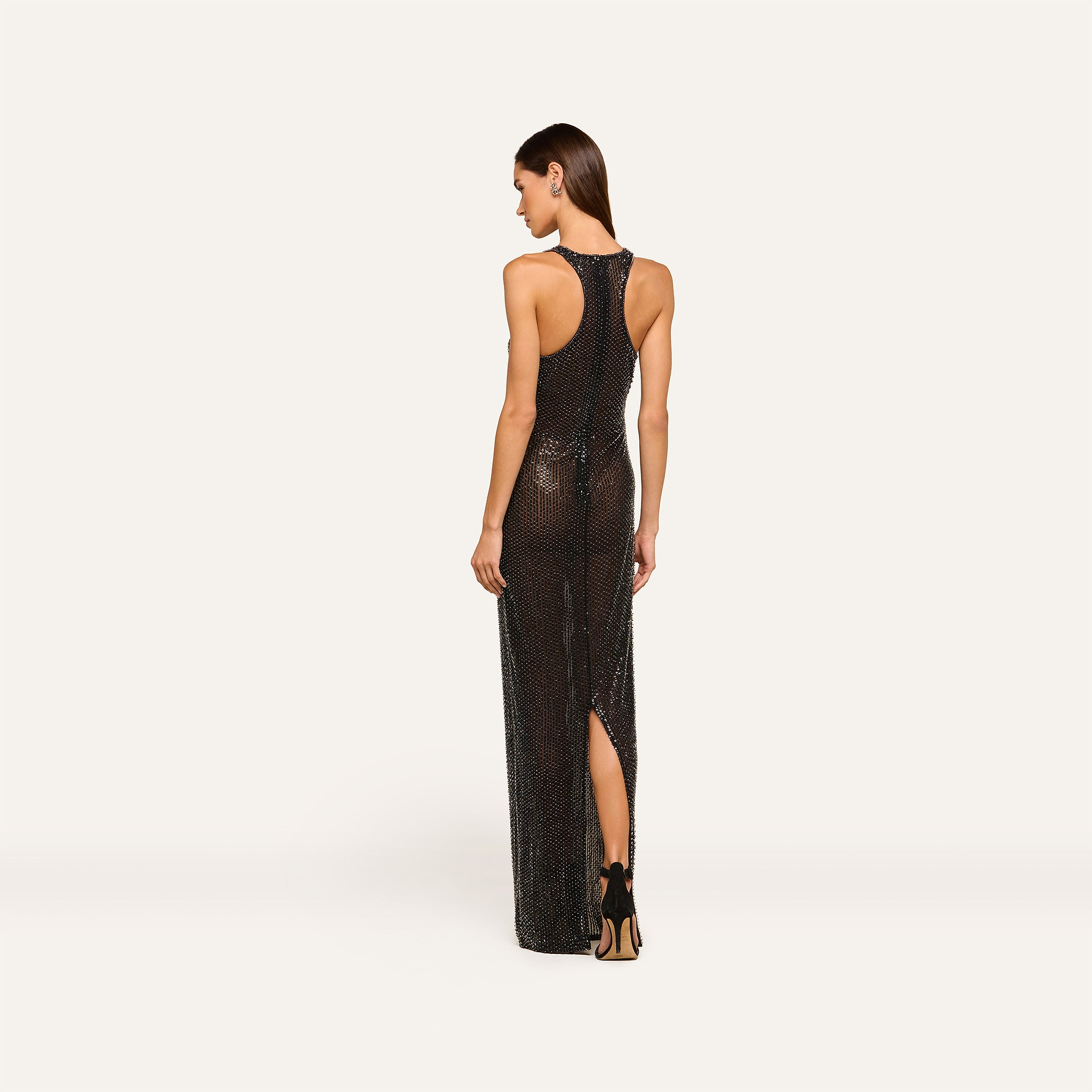 A woman stands with her back to the camera, showcasing the Paula Gown—a sleeveless, black racerback evening dress featuring a high slit—against a plain light background.