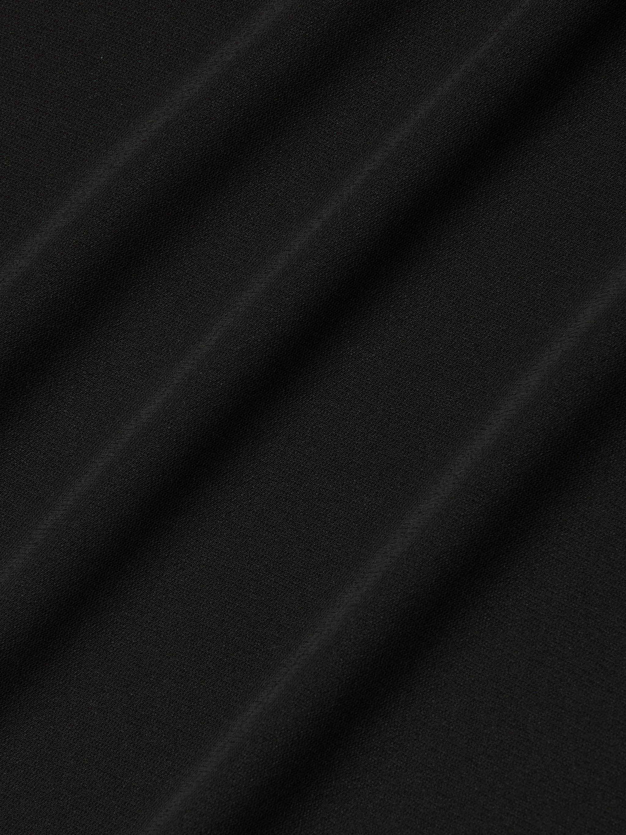Close-up of the Raye Slip Lining Dress in black stretch jersey fabric, featuring diagonal, evenly spaced folds that create a soft and subtle textured pattern. The surface appears smooth and matte, with shadows accentuating the gentle contours of the dress’s pleats.