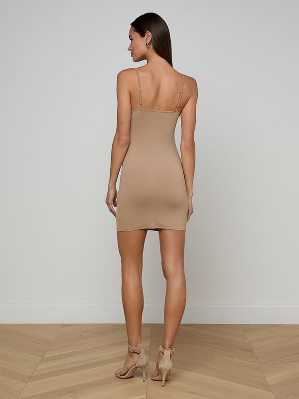 A woman stands with her back to the camera in a plain white room, wearing the beige Raye Slip Lining Dress—a sleeveless, stretch jersey fitted dress with adjustable straps—paired with high-heeled sandals.