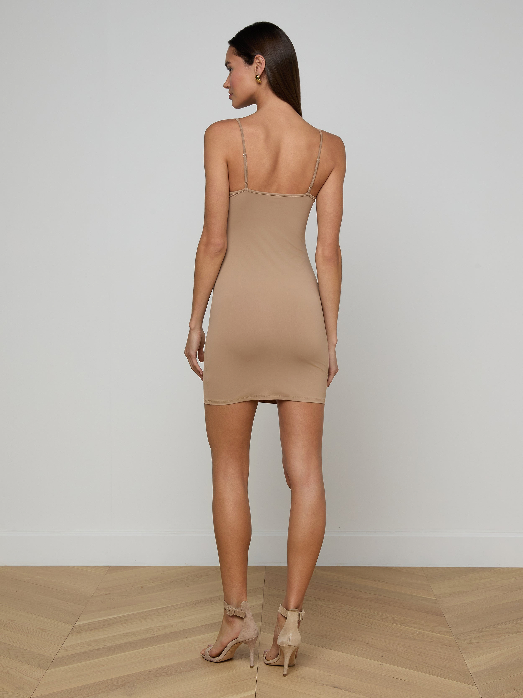 A woman stands with her back to the camera in a plain white room, wearing the beige Raye Slip Lining Dress—a sleeveless, stretch jersey fitted dress with adjustable straps—paired with high-heeled sandals.