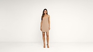 A woman stands in a plain, white room wearing a simple, beige sleeveless mini dress and beige heels, looking directly at the camera.