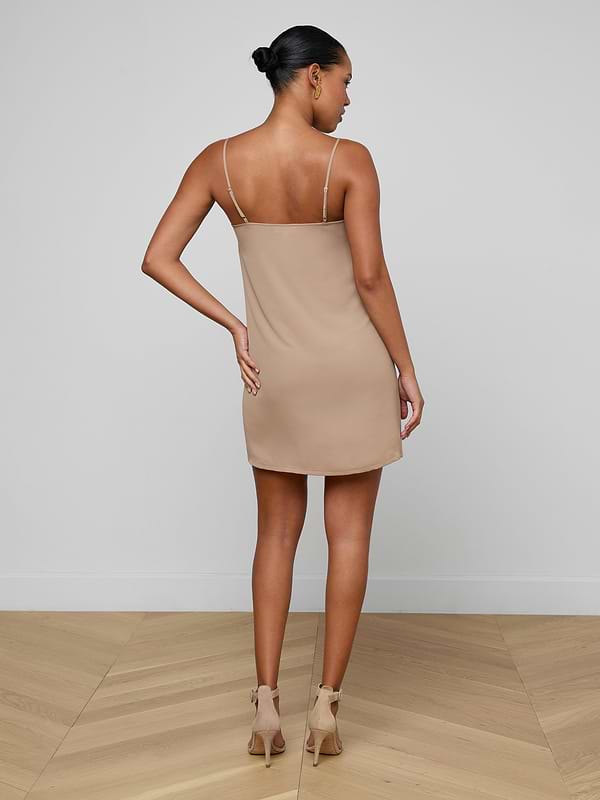 A woman stands facing away, wearing the Raye Slip Lining Dress—a beige, sleeveless mini dress featuring adjustable straps—paired with beige high-heeled sandals against a plain white background.