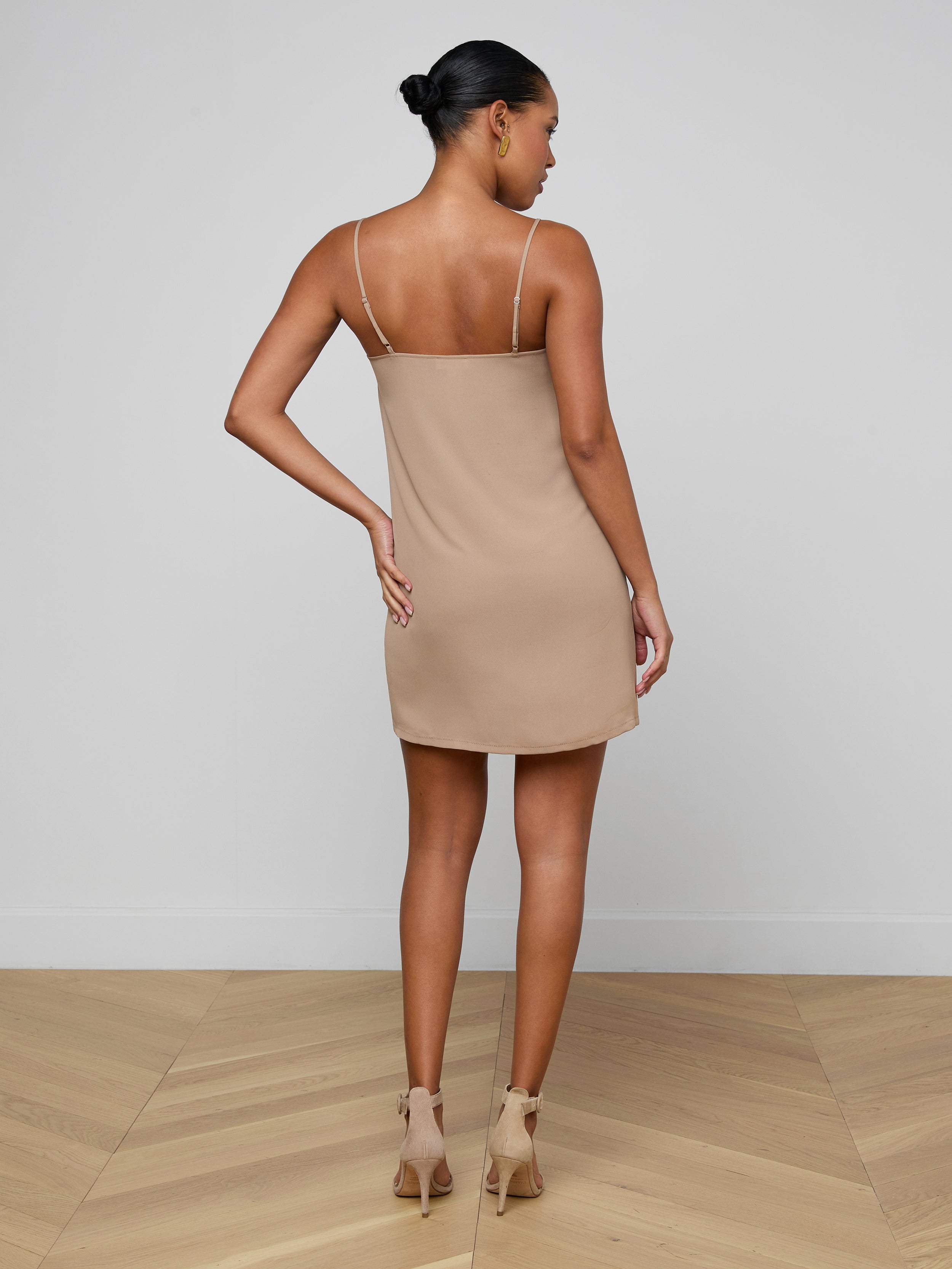 A woman stands facing away, wearing the Raye Slip Lining Dress—a beige, sleeveless mini dress featuring adjustable straps—paired with beige high-heeled sandals against a plain white background.