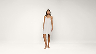 A woman stands in a white sleeveless dress and white sandals against a plain white background.