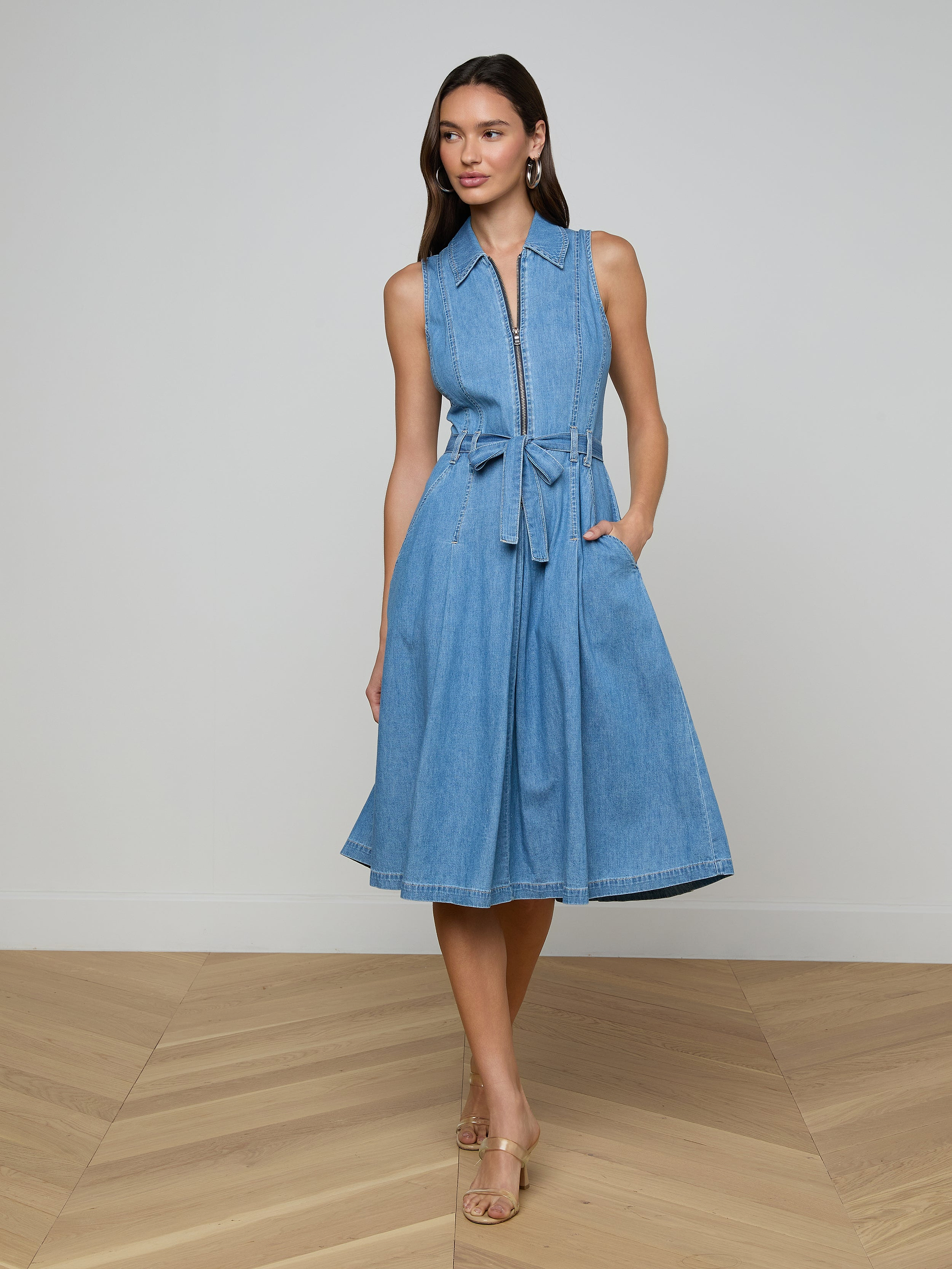 A woman wears the Kieran Shirt Dress—a sleeveless, knee-length, featherweight denim style with a tie belt and collar—paired with beige heels. She stands on a wooden floor against a plain light wall, one hand in her pocket.