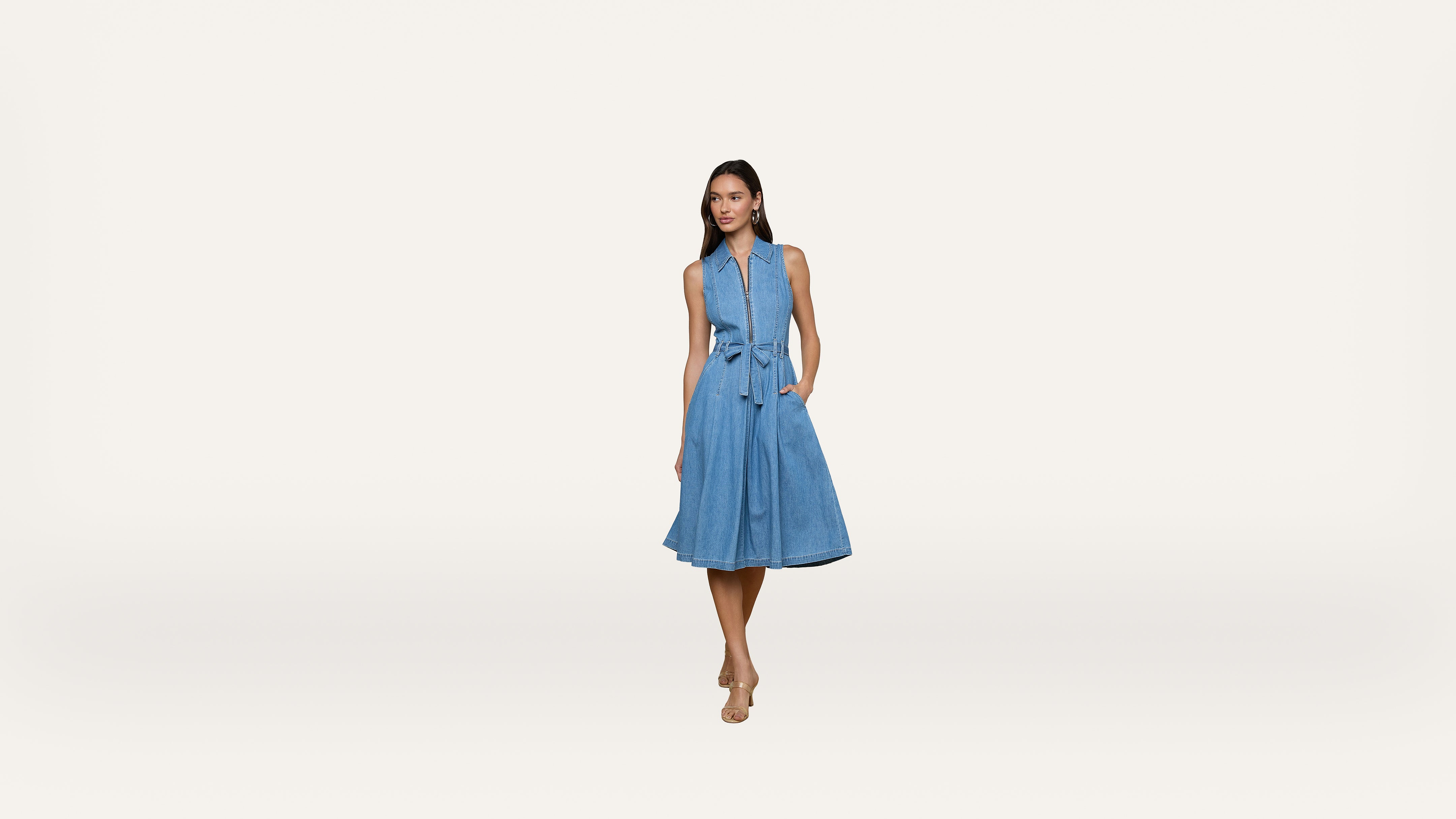 A woman stands against a plain white background, wearing a sleeveless blue denim dress with a tied waist and tan heels.