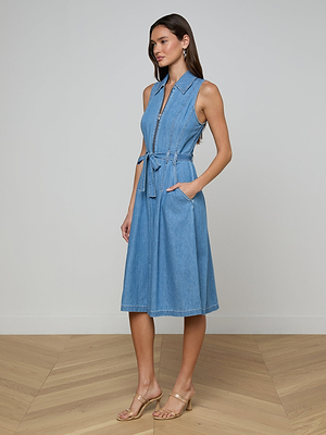 A woman stands on a wooden floor wearing the Kieran Jean Shirt Dress—a sleeveless, light blue featherweight denim dress with a collar, front zipper, tie belt, and pockets. She pairs it with beige heeled sandals and has long hair.