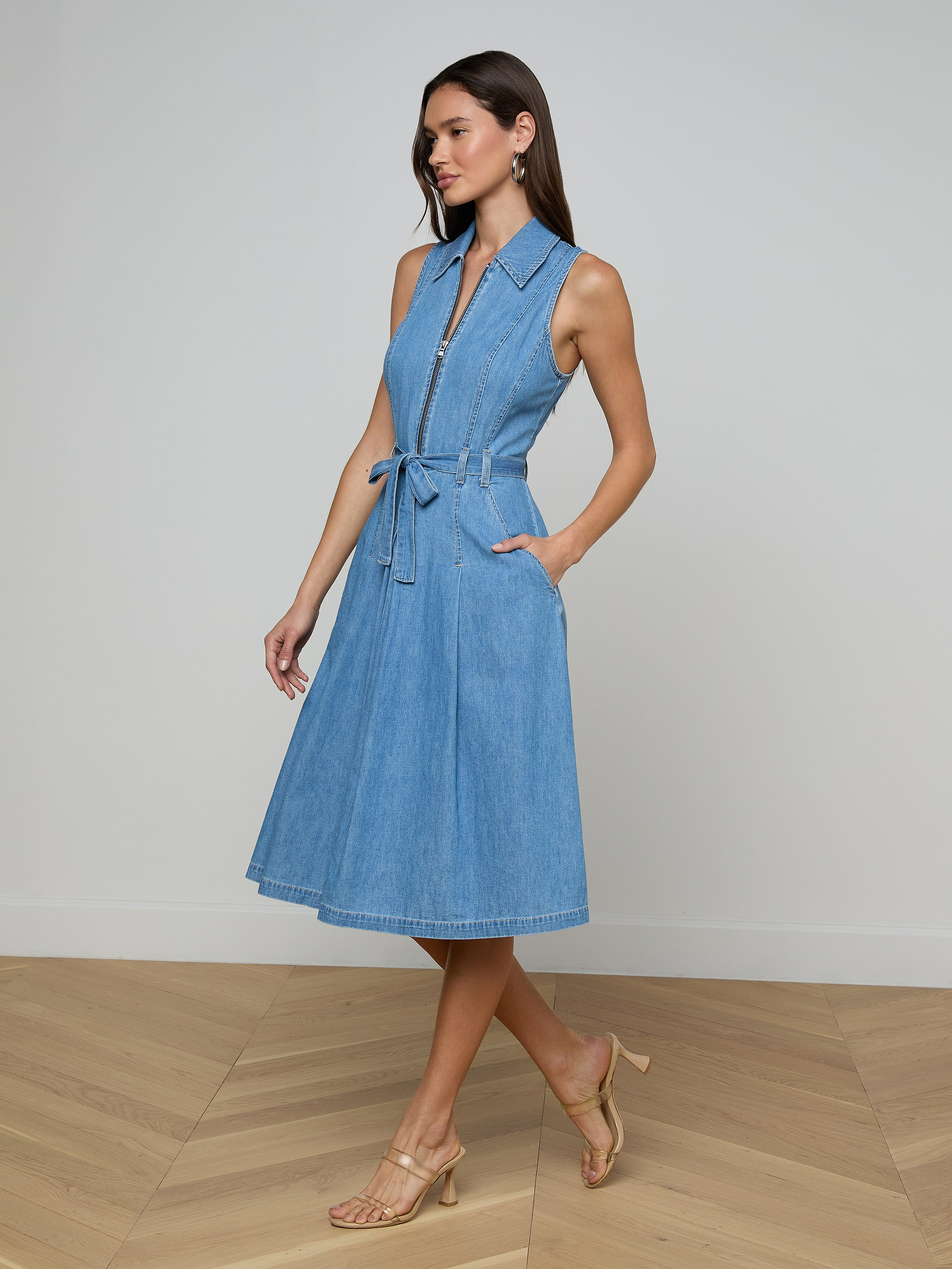 A woman in a minimalist room wears the Kieran Shirt Dress, featuring a collar, front zipper, tie belt, and side pockets. She completes the look with nude heeled sandals and has long, straight hair.