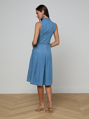A woman stands on a wooden floor facing away, wearing the sleeveless, knee-length Kieran Jean Shirt Dress in blue featherweight denim with a tie belt and beige heeled sandals; her hair is down and hands relaxed by her side.