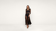 A woman stands against a plain white background wearing a long, sheer black lace dress and black high-heeled sandals.