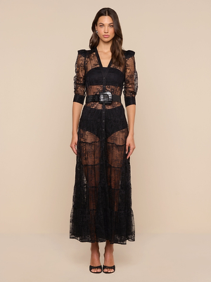Seraphina wears the Seraphina Lace Tiered Maxi Dress, a sheer black lace design with mid-length sleeves, deep V-neck, black belt, visible undergarments, and black open-toe heels against a beige background.