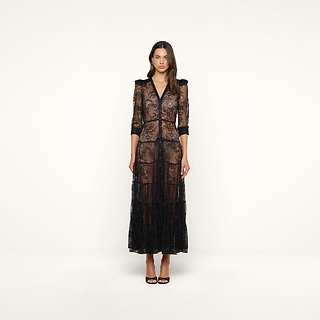 A woman stands facing forward against a plain white background, wearing the Seraphina Lace Tiered Maxi Dress—a sheer black lace bohemian maxi dress featuring 3/4 length sleeves and a V-neckline—paired with black heels.