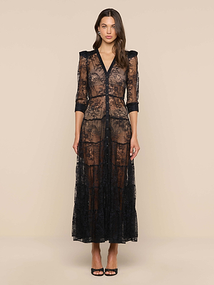 A woman with long, wavy brown hair faces forward in the Seraphina Lace Tiered Maxi Dress—sheer lace with three-quarter sleeves, a V-neckline, and black trim. She pairs it with black open-toe heels against a beige background.
