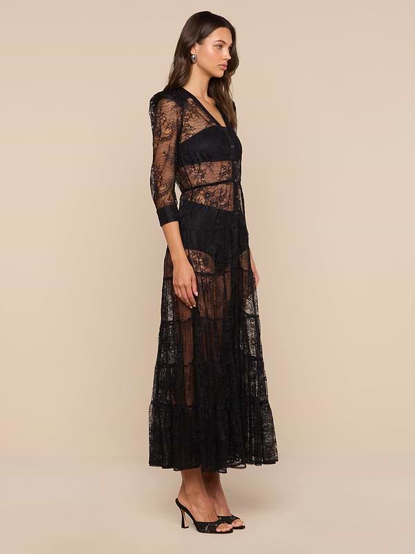 A woman stands sideways against a plain white background, wearing the Seraphina Lace Tiered Maxi Dress—a sheer, long black bohemian maxi dress with 3/4 length sleeves—paired with black heels.