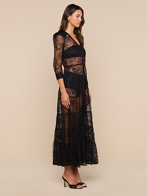Woman standing sideways in a Seraphina Lace Tiered Maxi Dress with three-quarter sleeves, sheer lace detailing, black undergarment set, and black heeled sandals against a plain beige background.