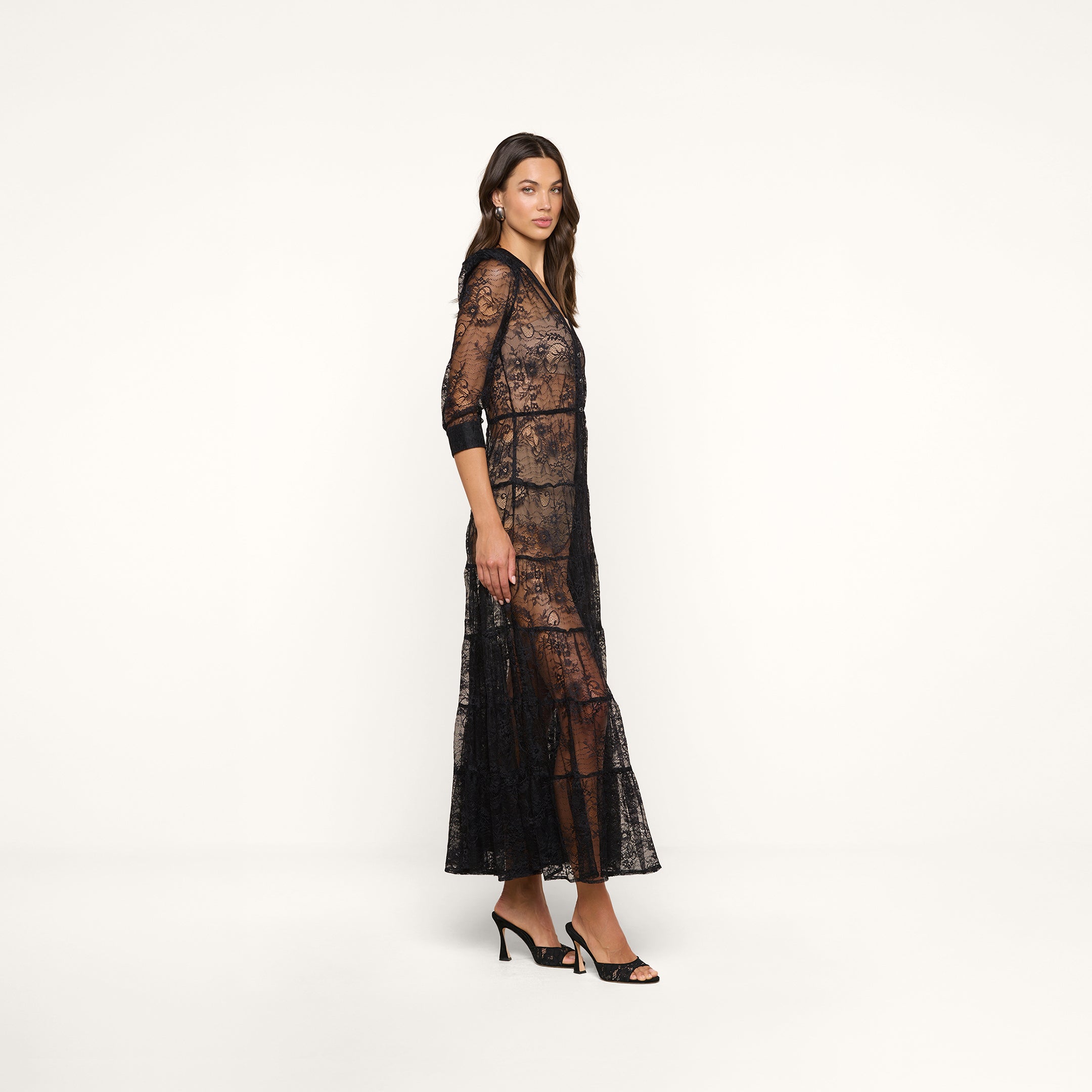 A woman stands against a plain white background, wearing the Seraphina Lace Tiered Maxi Dress—a sheer lace black bohemian style with 3/4 length sleeves—paired with black high-heeled sandals. She faces slightly to the side, her expression neutral.
