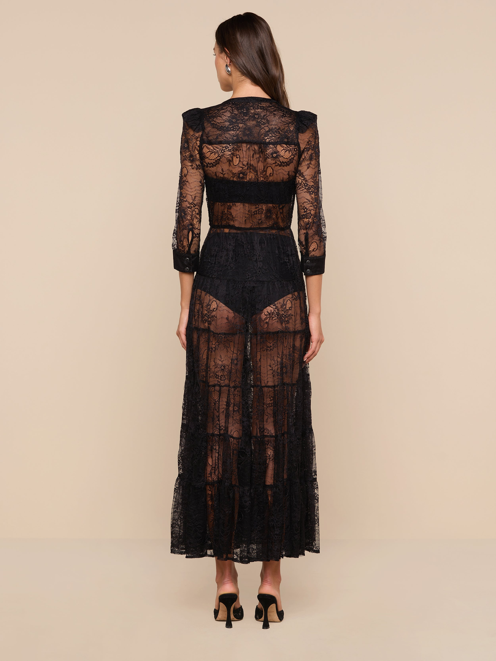 A woman stands facing away, wearing the Seraphina Lace Tiered Maxi Dress—a sheer, bohemian-style lace maxi dress with 3/4 length sleeves and visible undergarments. Her long brown hair cascades down her back, and she completes the look with black high-heeled shoes against a plain white background.