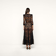 A woman with long brown hair, wearing the Seraphina Lace Tiered Maxi Dress—sheer black lace with 3/4 length sleeves—paired with black heels, stands facing away against a plain light background.