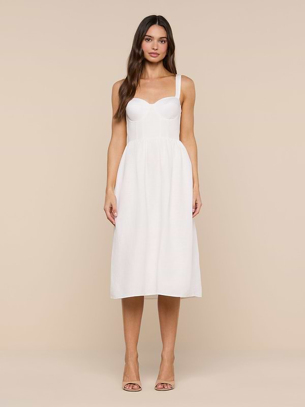 A woman stands facing forward wearing the Tiani Bustier Dress—a white, sleeveless, knee-length linen dress featuring a fitted bodice and delicate spaghetti straps. She has long, loose hair and is styled with nude heels against a plain white background.