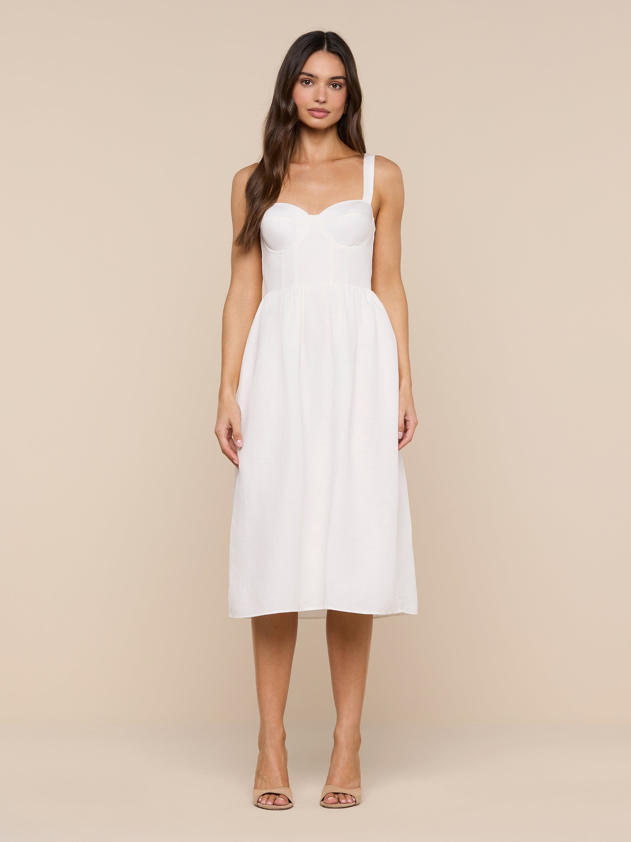 A woman stands facing forward wearing the Tiani Bustier Dress—a white, sleeveless, knee-length linen dress featuring a fitted bodice and delicate spaghetti straps. She has long, loose hair and is styled with nude heels against a plain white background.