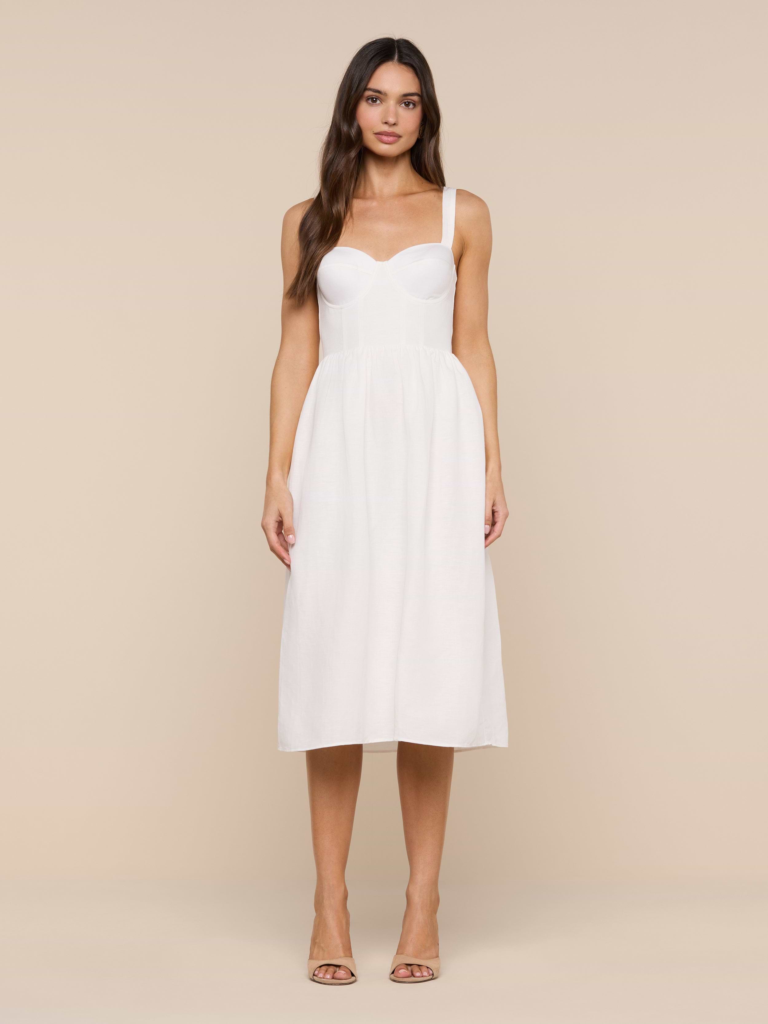 A woman stands facing forward wearing the Tiani Bustier Dress—a white, sleeveless, knee-length linen dress featuring a fitted bodice and delicate spaghetti straps. She has long, loose hair and is styled with nude heels against a plain white background.