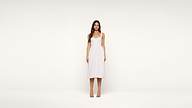 Woman stands facing forward, wearing a knee-length white dress with thin straps and nude heels, against a plain white background.