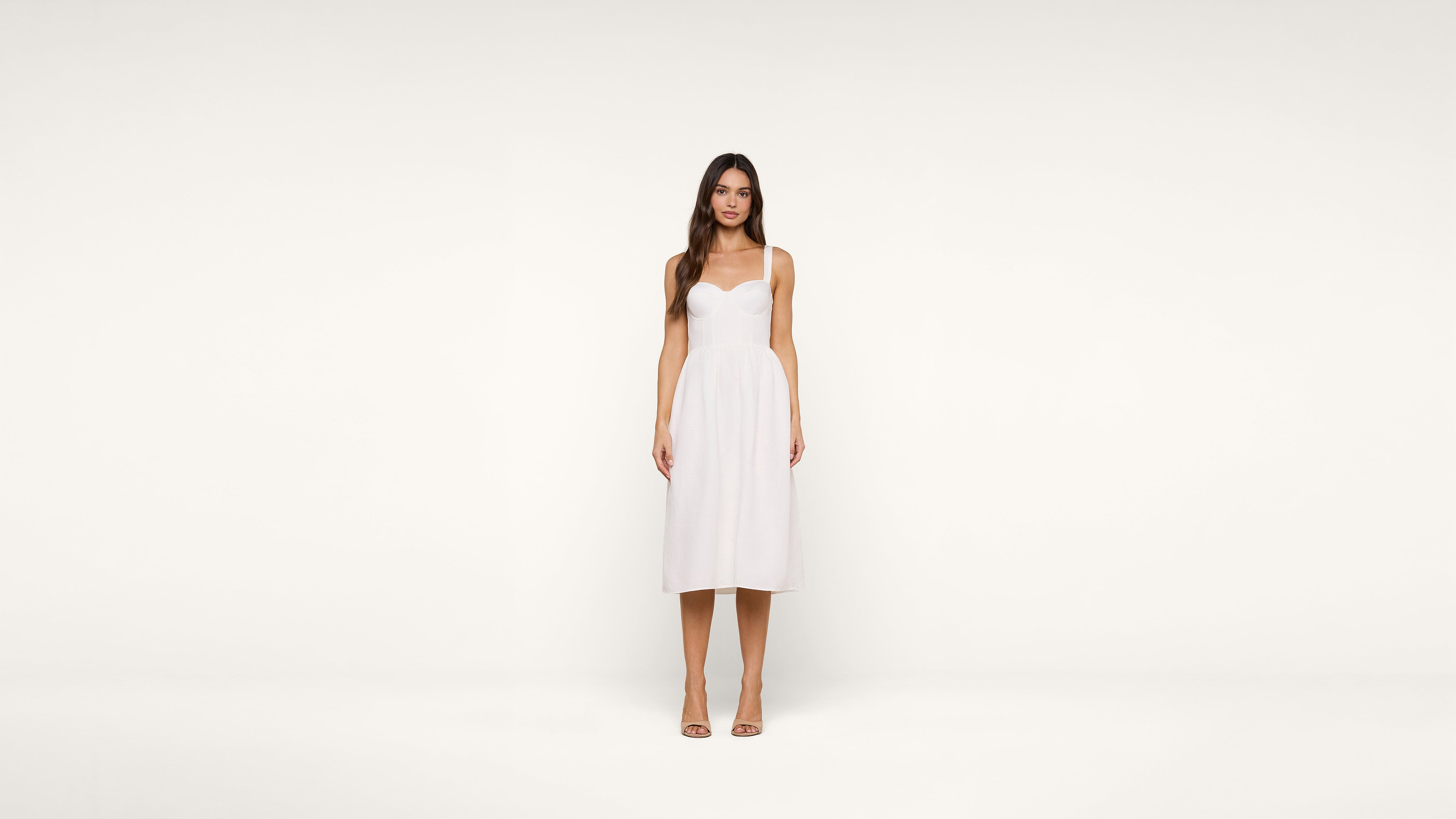 Woman stands facing forward, wearing a knee-length white dress with thin straps and nude heels, against a plain white background.