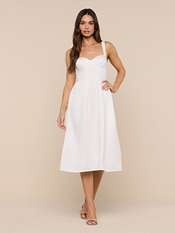A woman with long brown hair and a neutral expression stands against a white background, wearing the Tiani Bustier Dress—a sleeveless, fitted white linen summer dress with thin straps—paired with tan heeled sandals.