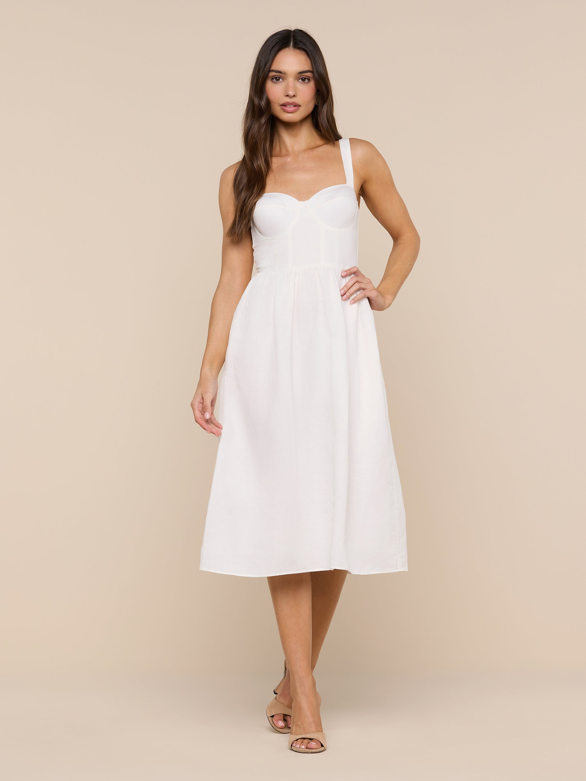 A woman with long brown hair and a neutral expression stands against a white background, wearing the Tiani Bustier Dress—a sleeveless, fitted white linen summer dress with thin straps—paired with tan heeled sandals.