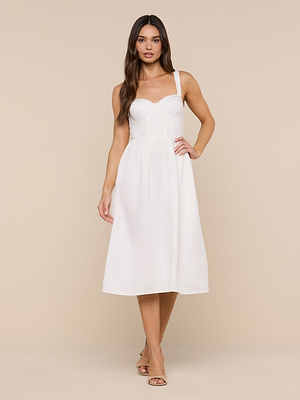 A woman wears the Tiani Bustier Dress—a white, sleeveless linen dress with a fitted bodice and flared skirt—standing against a beige background in nude heels; an ideal summer look.