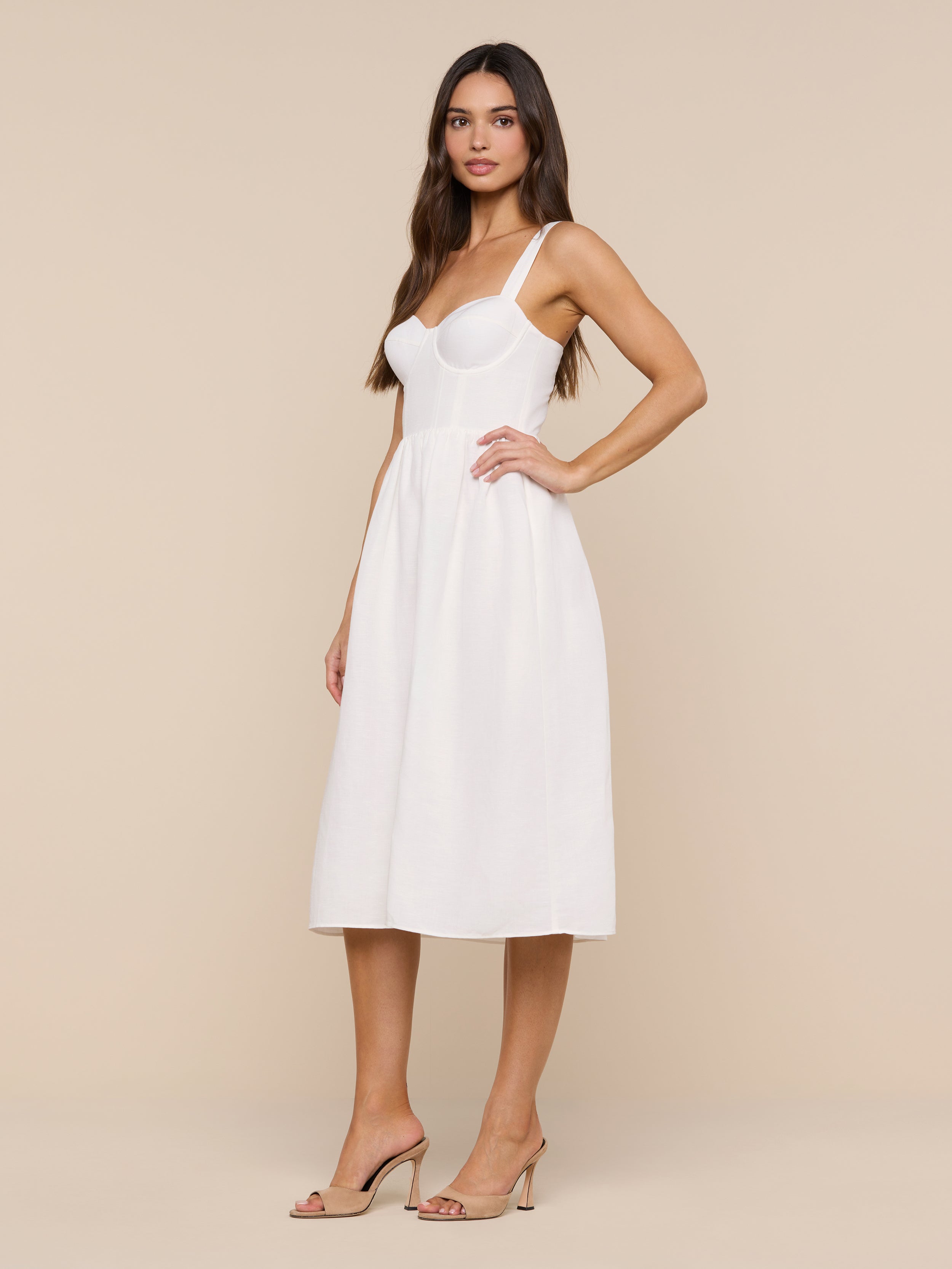 A woman with long brown hair poses with her hand on her hip against a plain white background, wearing the Tiani Bustier Dress—a white linen summer dress featuring underwire cups—paired with nude heels.