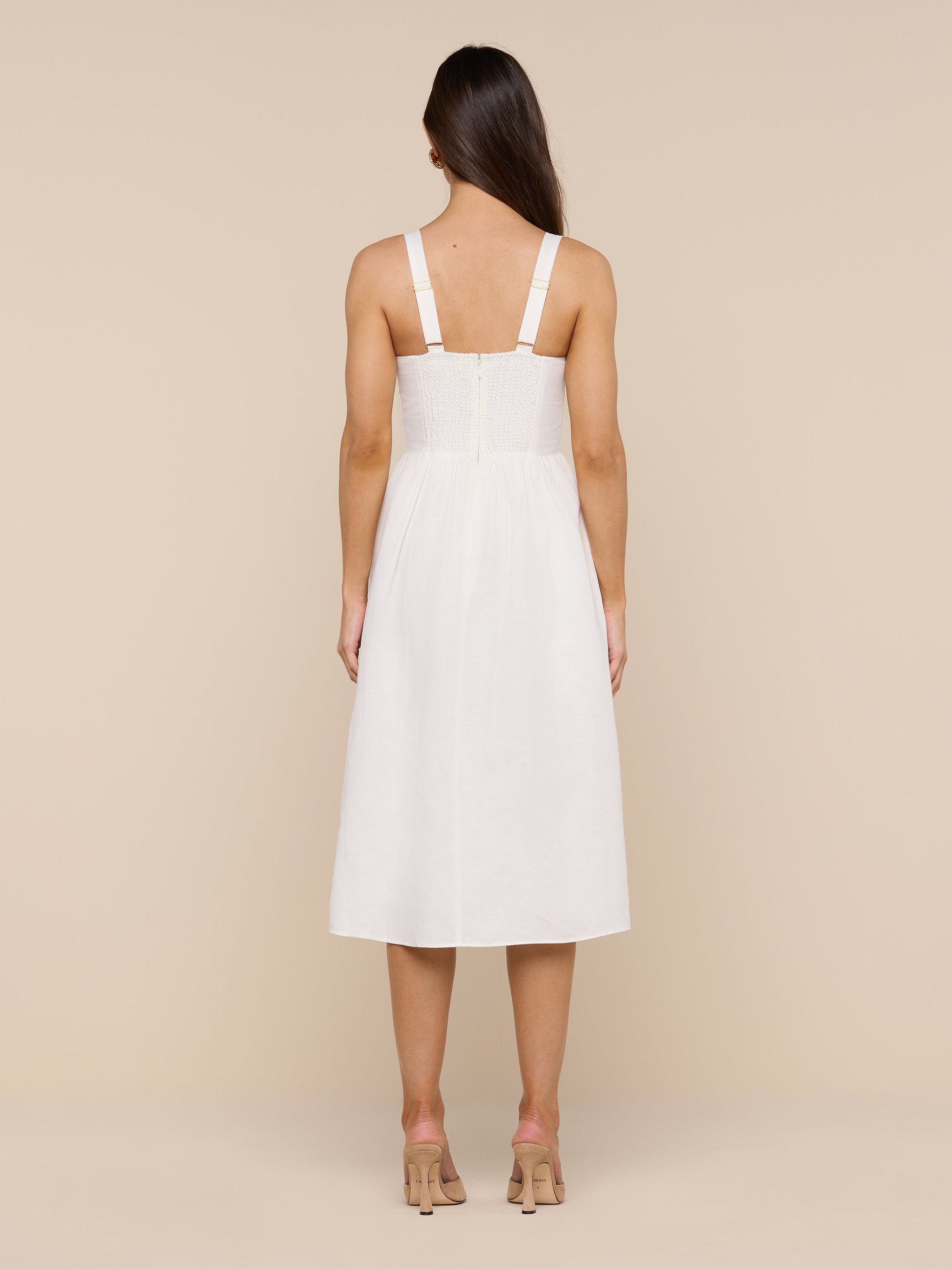 A woman stands facing away, wearing the Tiani Bustier Dress—a white, sleeveless, knee-length linen dress with wide straps—and nude heels against a plain white background.