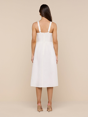 A woman with long brown hair faces away, wearing the sleeveless Tiani Bustier Dress in white linen with thick straps and beige high heels against a neutral background.