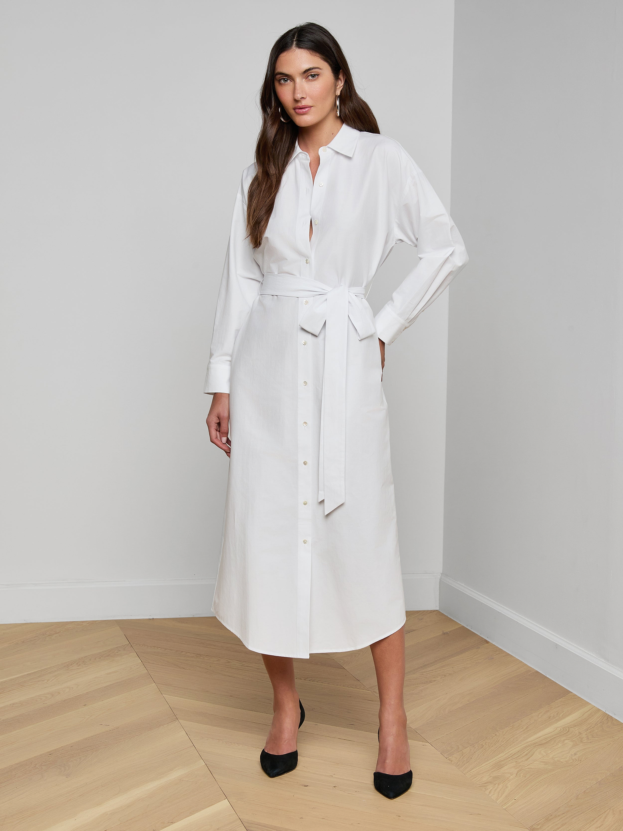 The woman is wearing the Ridley Shirt Dress, a long-sleeved, oversized white dress crafted from organic cotton, complete with a matching waist tie. She stands against a plain white background with one hand on her hip and pairs the look with black heels.